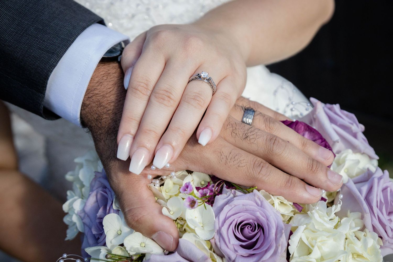 Newlyweds display wedding rings and diamond engagement ring over a purple and white floral bouquet.