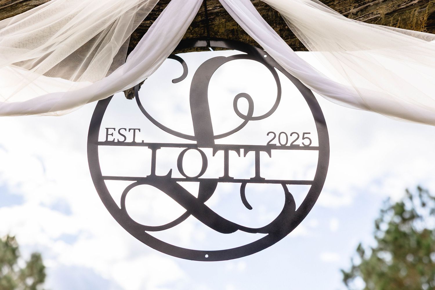 Decorative black metal Lott family monogram sign established 2025 hanging under a draped arbor.