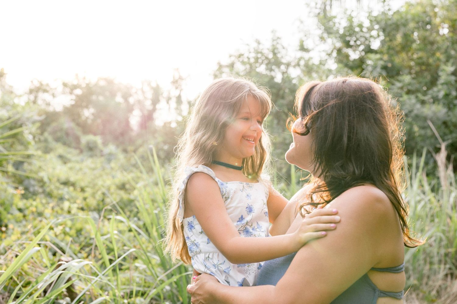 A mother and young daughter share a tender moment outdoors in a sunlit green meadow.