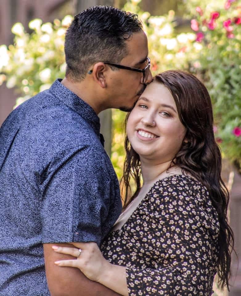 A couple shares a tender moment outdoors, man kissing woman's forehead while she smiles, with flowers in background.