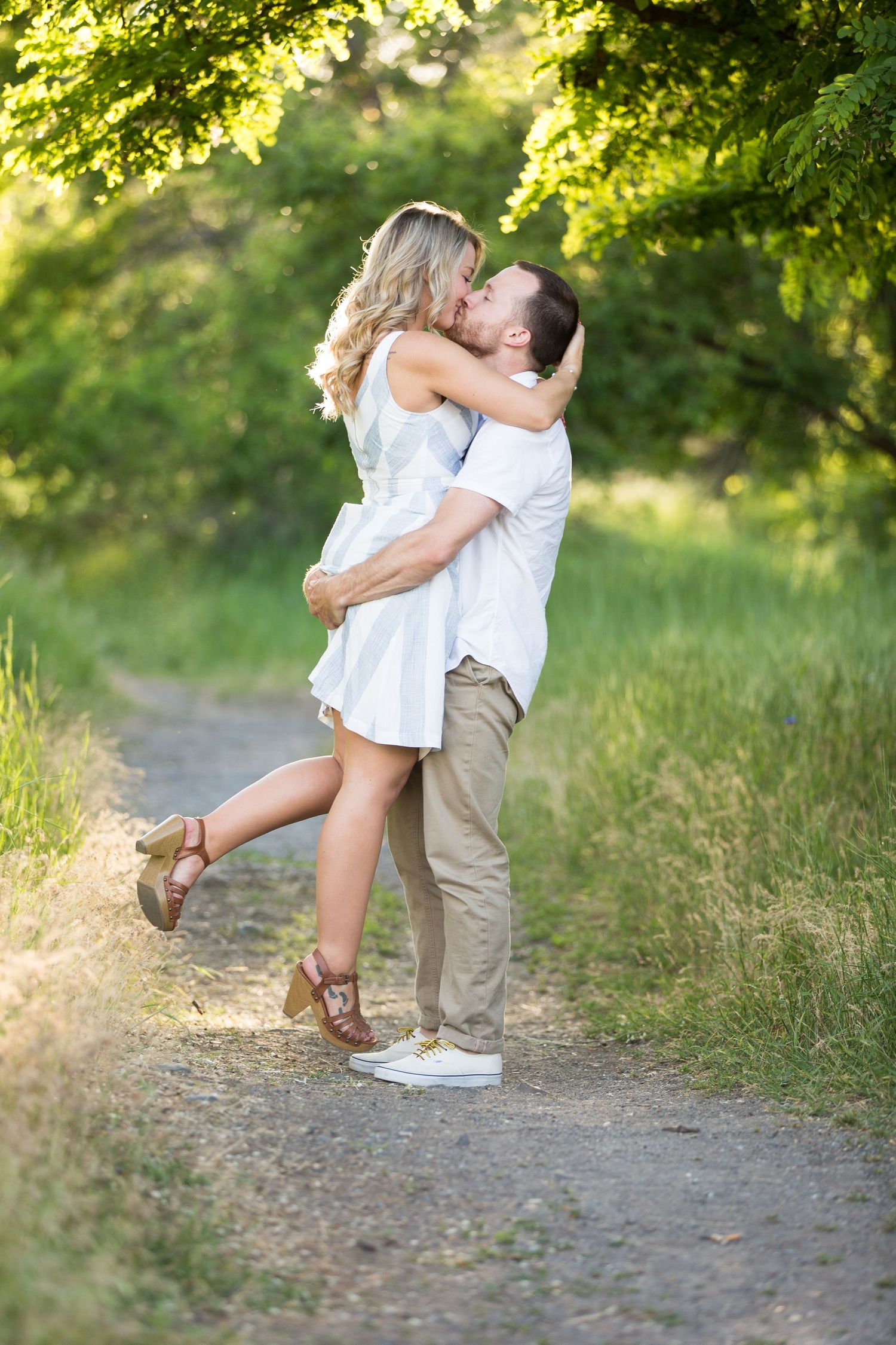A joyful couple kisses on a sunlit path, the man lifting the woman in a white dress off the ground.