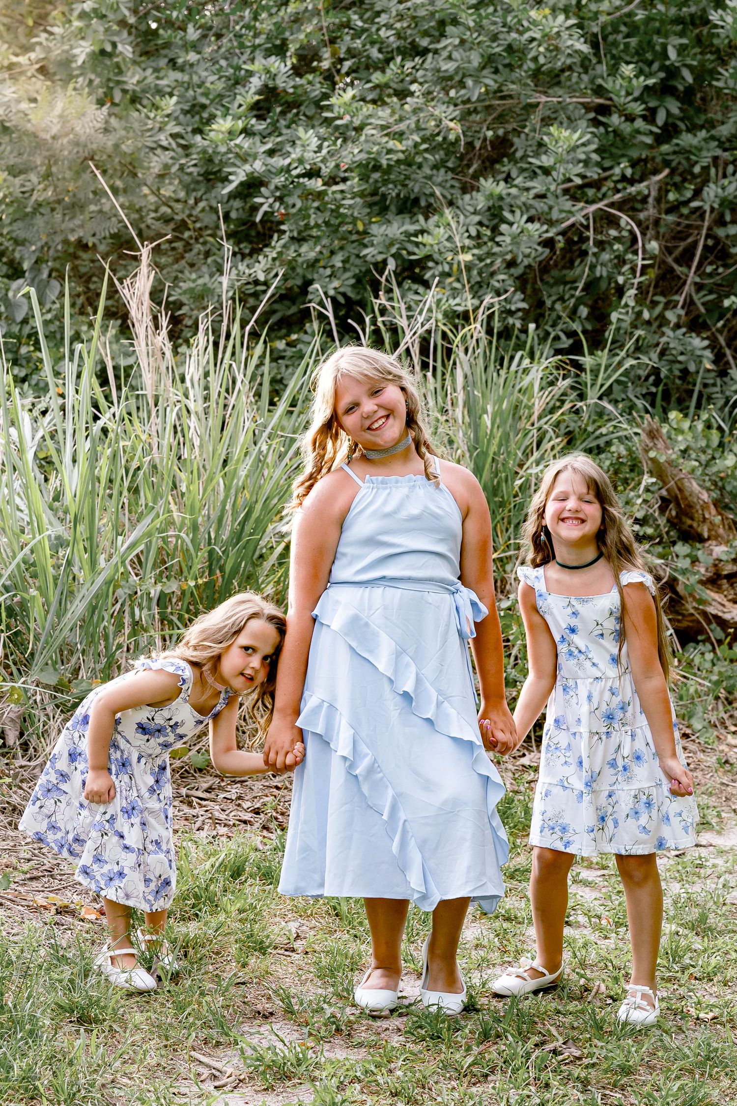 Three girls in blue floral dresses hold hands and smile outdoors surrounded by lush green foliage.
