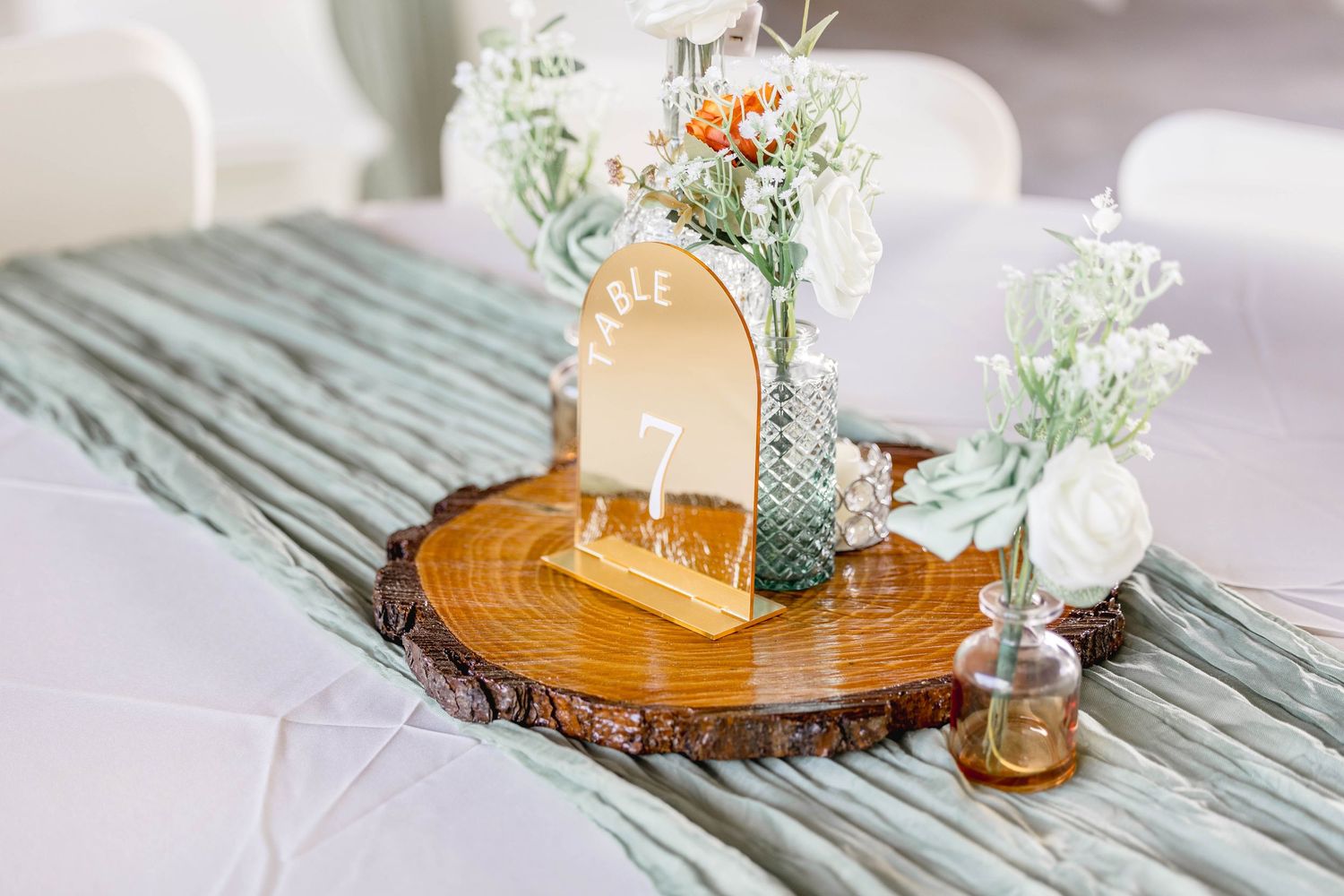 Elegant wedding table centerpiece with wood slice, gold table number, and white floral arrangements.