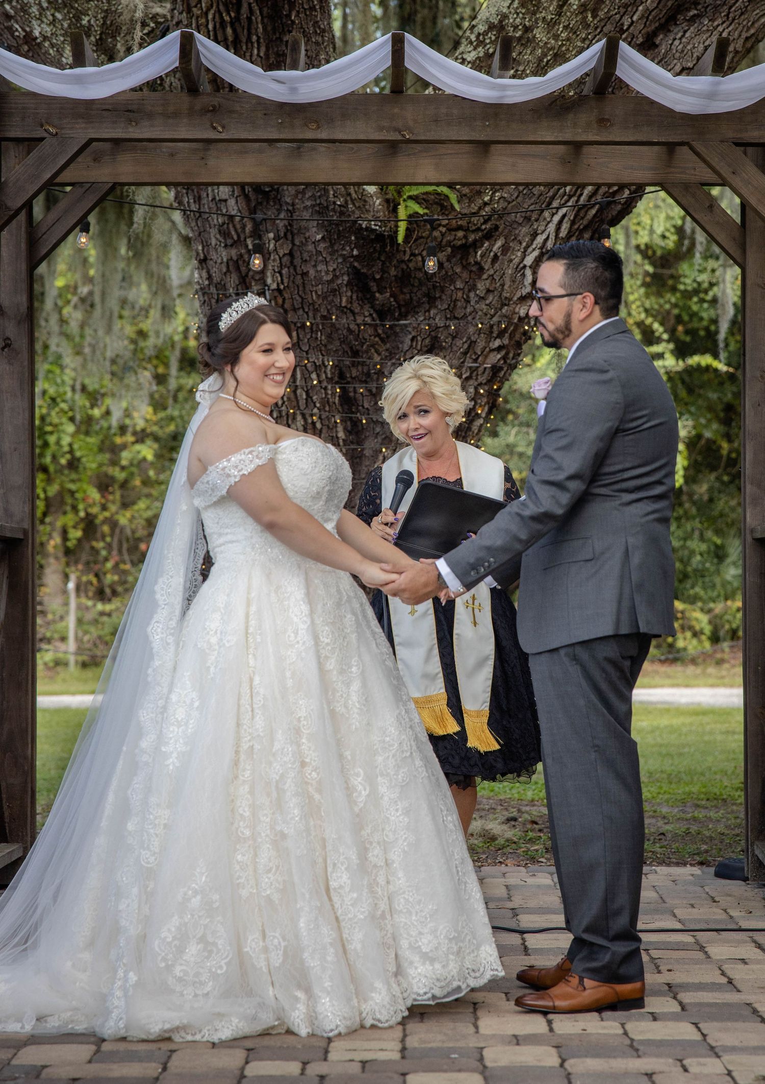 Bride and groom hold hands exchanging vows under rustic wooden pergola with officiant at outdoor wedding.