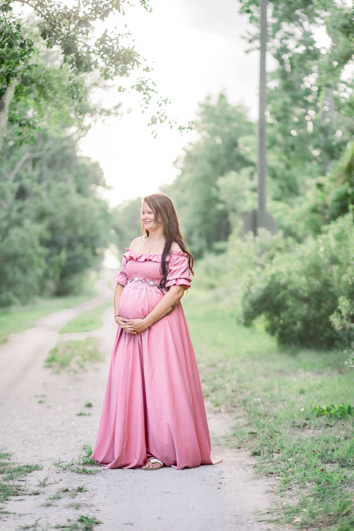 Pregnant woman in pink off-shoulder maternity gown standing on a serene tree-lined path.