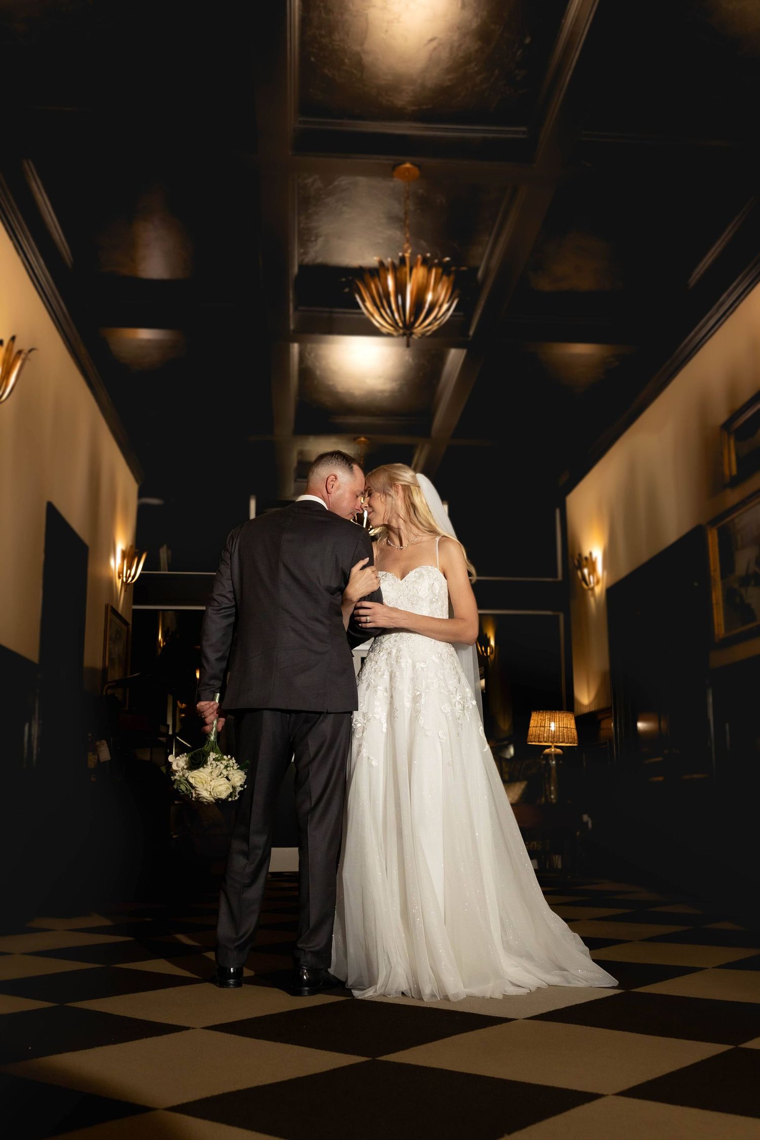 Bride and groom share intimate moment in dramatic dark hallway of elegant wedding venue.