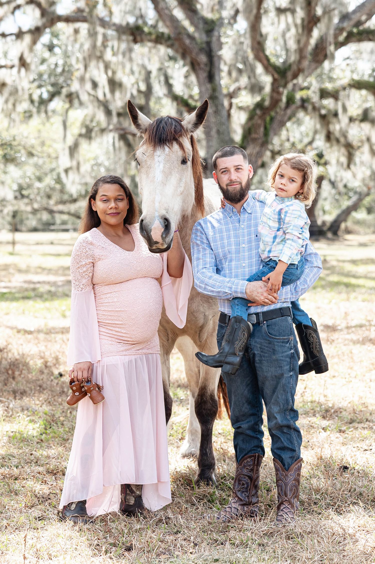 Pregnant woman in pink dress with family, child, and horse under moss-draped trees.