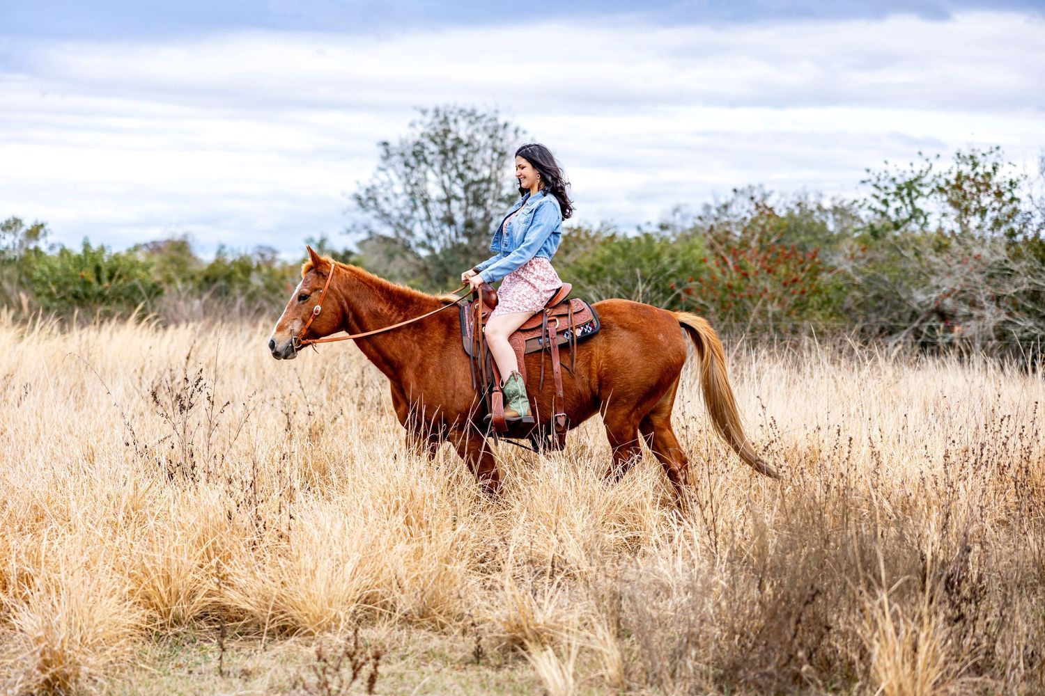 Young woman in denim jacket riding chestnut horse through golden dry grass field.