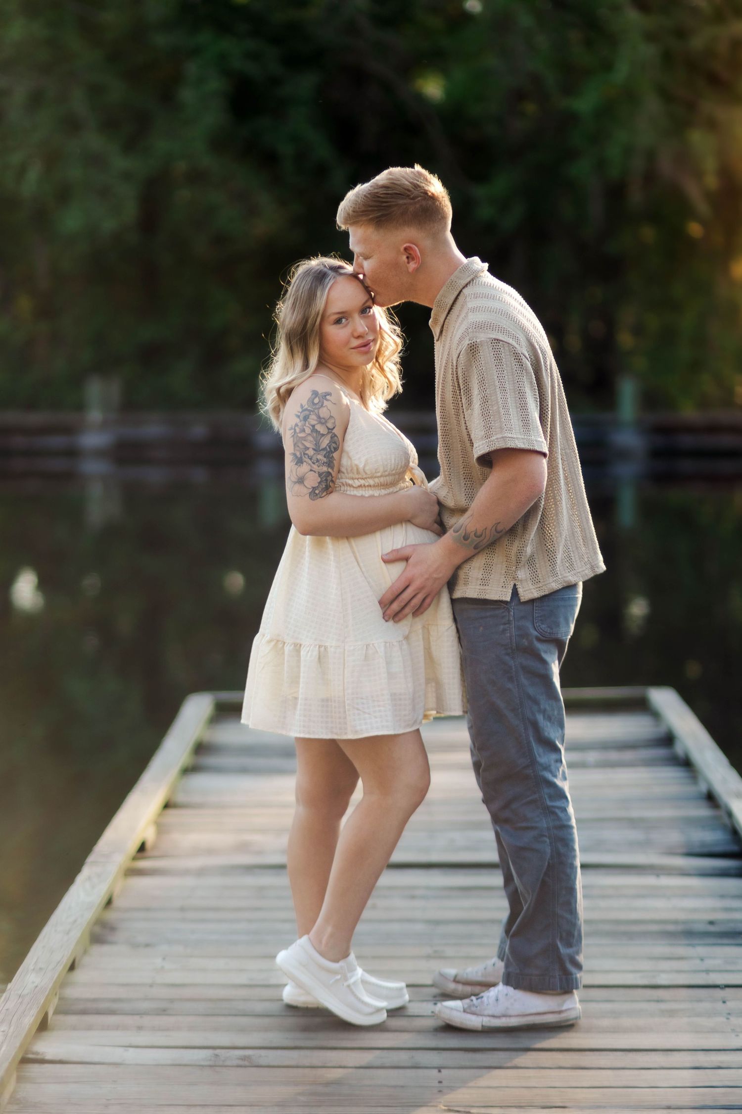 Pregnant couple embracing on wooden dock by lake, man kissing woman's head at sunset.