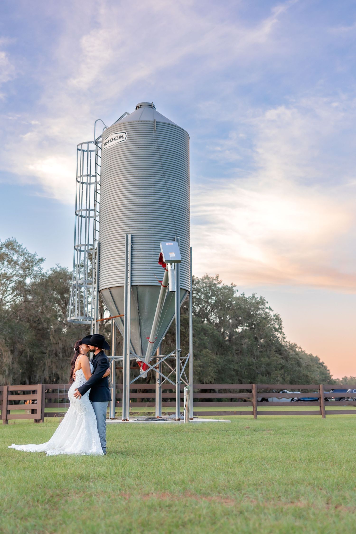 Bride and groom embrace near a farm silo at sunset during their rustic outdoor wedding ceremony.