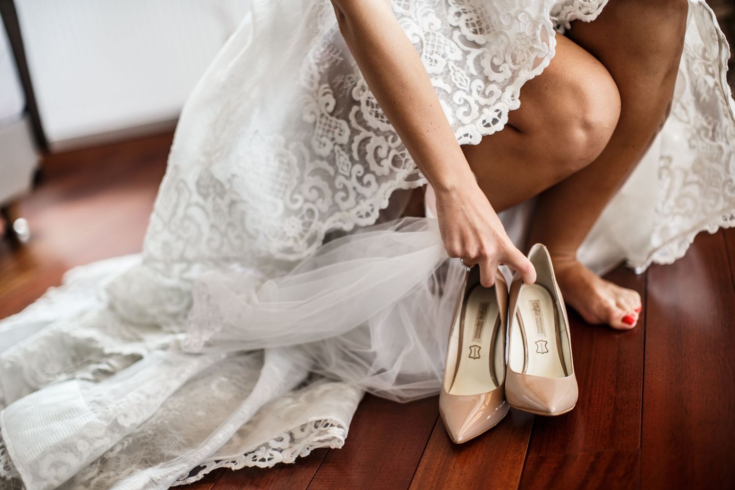 Bride in lace wedding dress putting on nude heels while sitting on wooden floor.