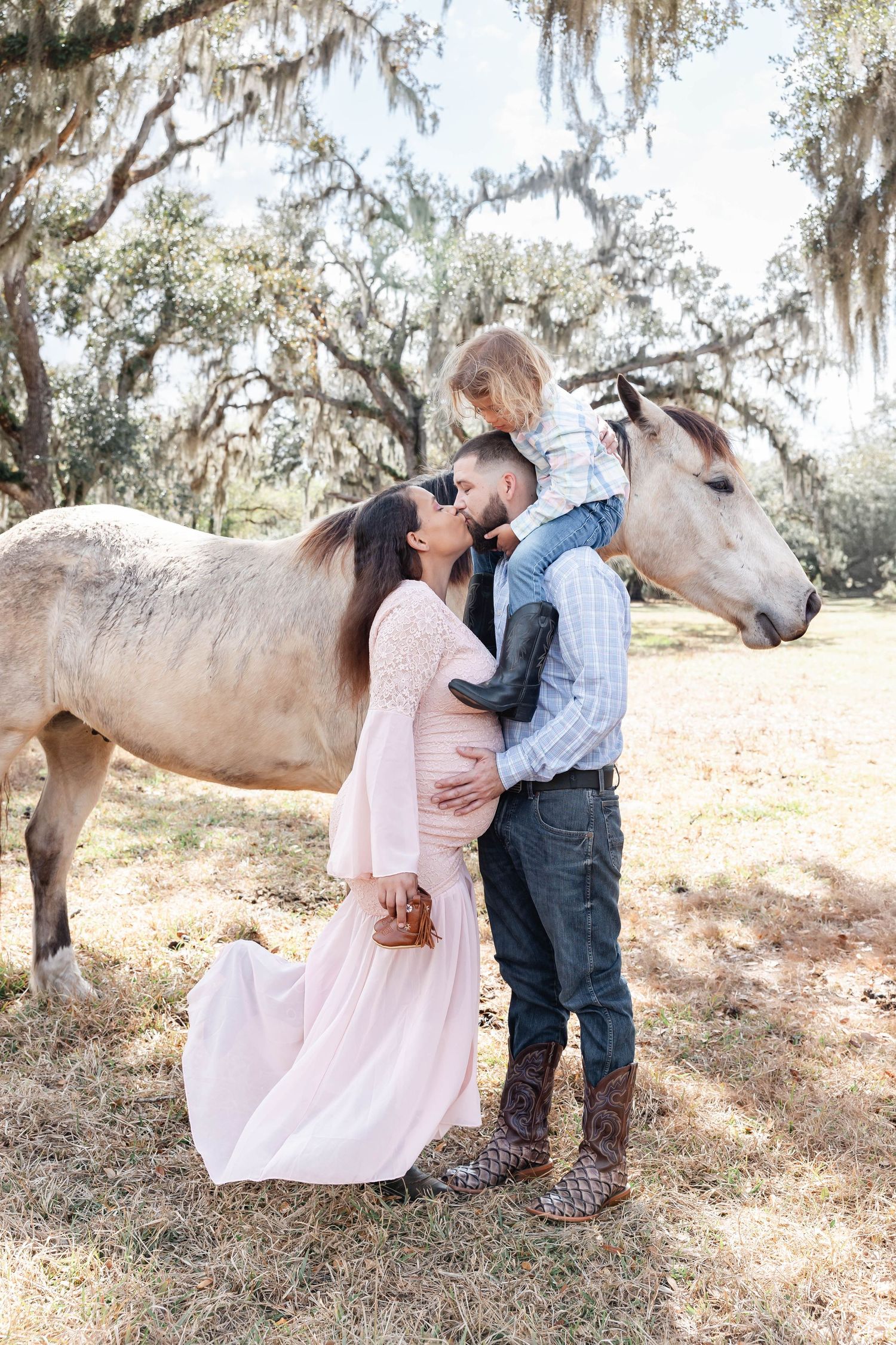 Couple sharing a kiss while man holds toddler, standing beside a gray horse in a sunlit mossy oak setting.