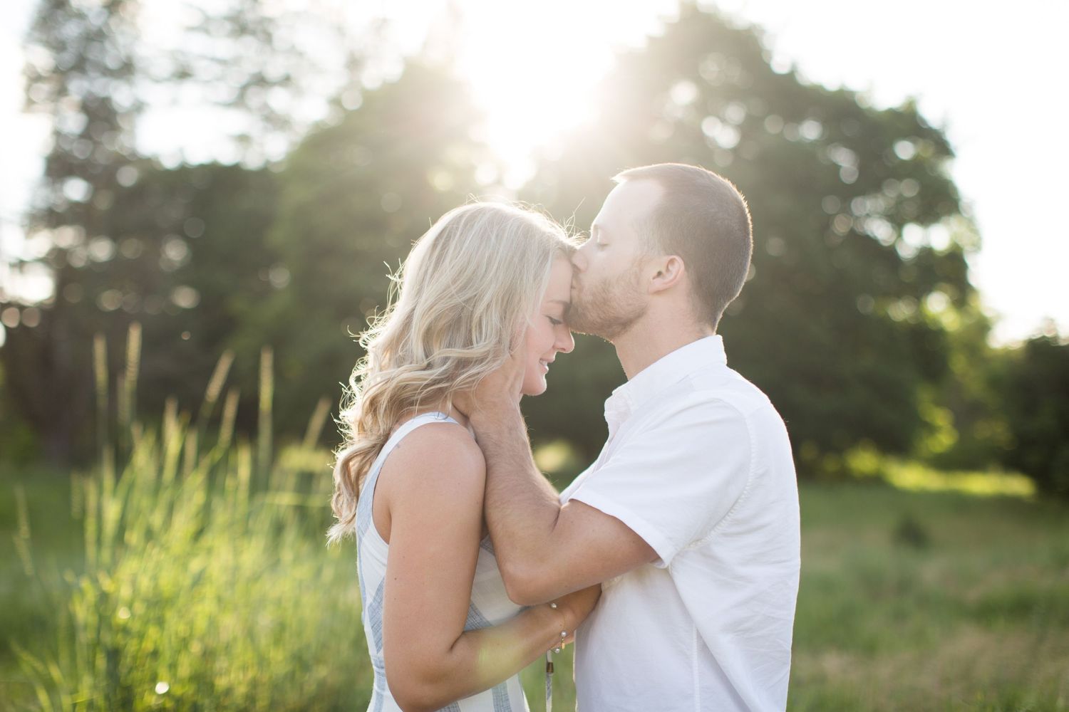 Couple embraces and touches foreheads in a sunny green field during golden hour engagement session.