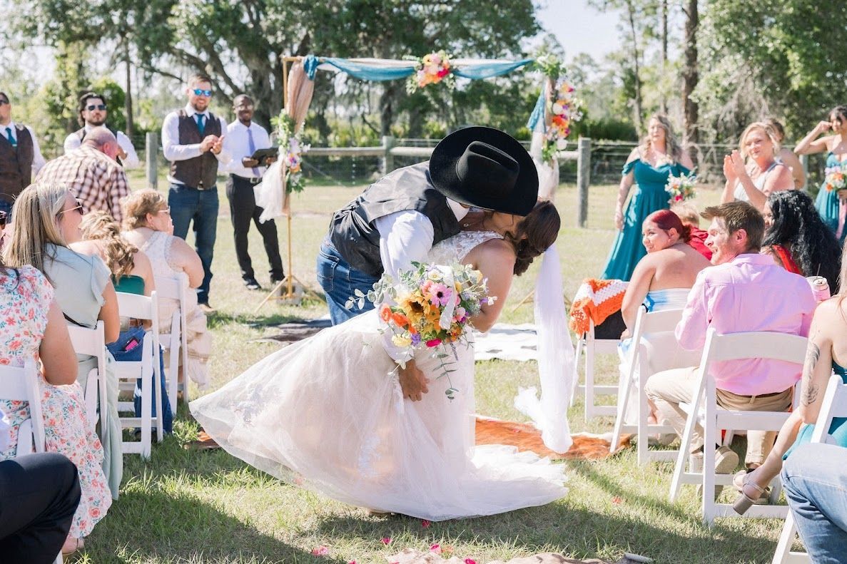 Groom dips bride for romantic kiss at outdoor wedding ceremony with guests watching.