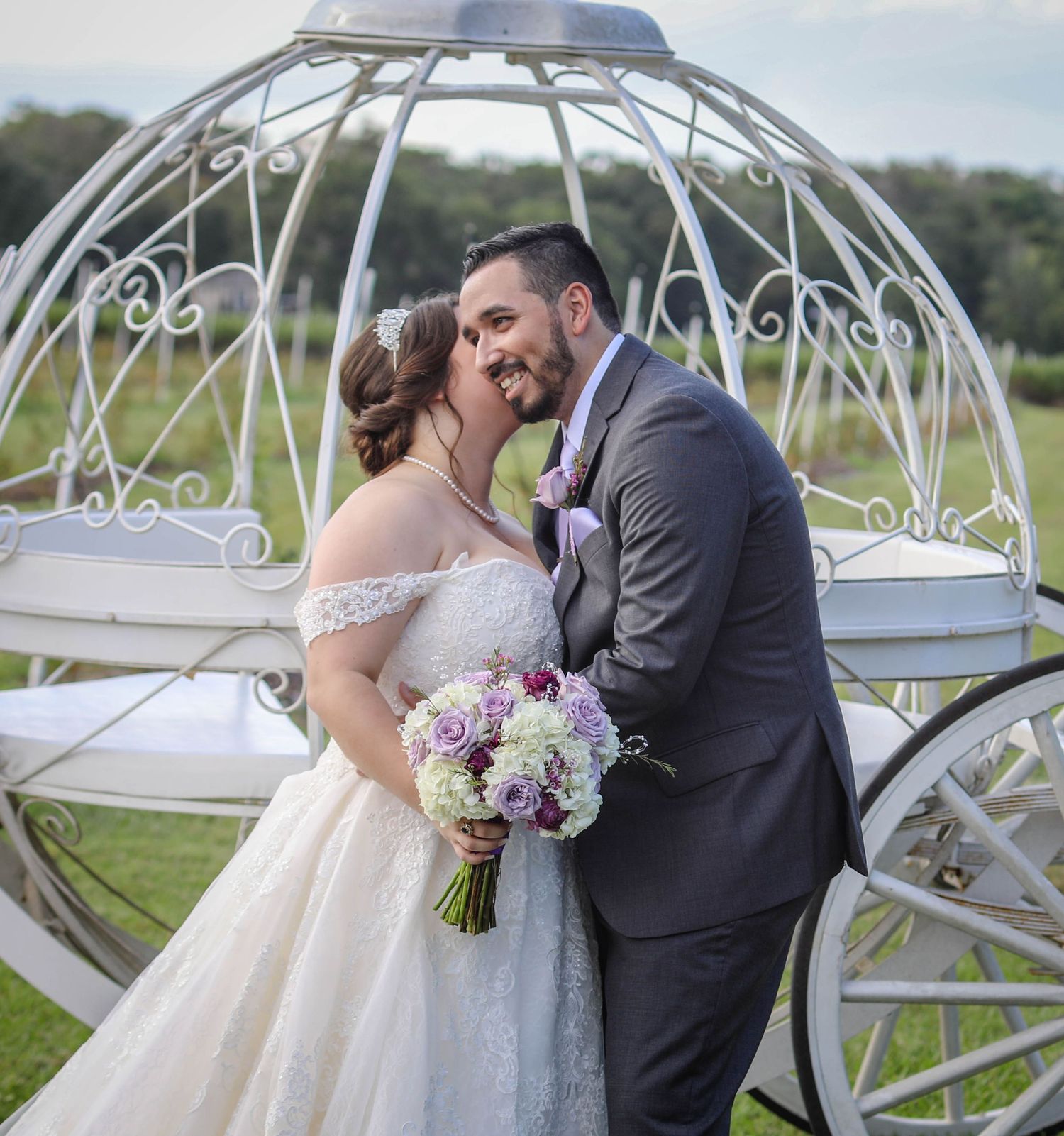 Bride and groom kissing beside white Cinderella carriage on green lawn with purple bridal bouquet.