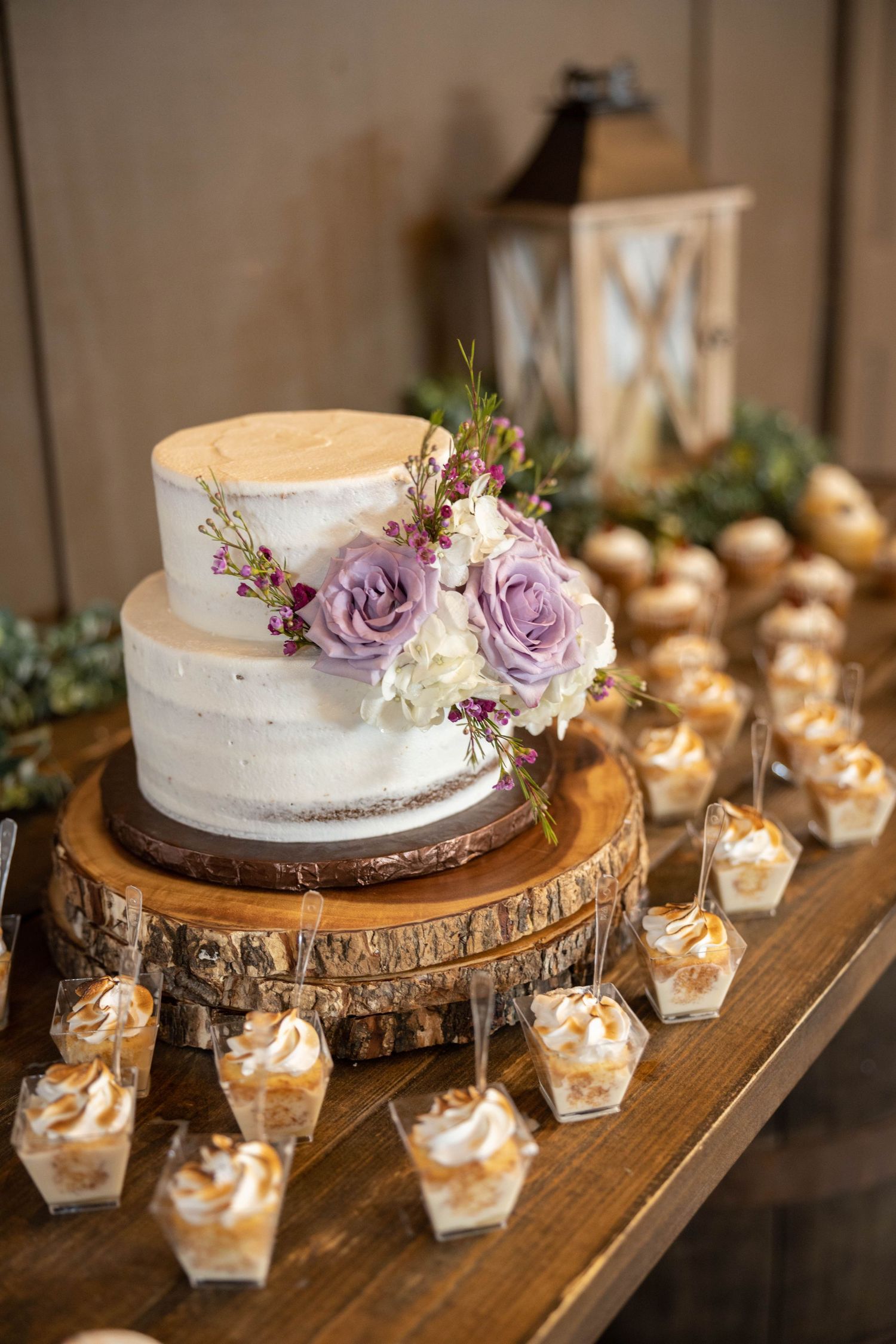 Two-tier white naked wedding cake with lavender roses on wood slice stand with dessert table.