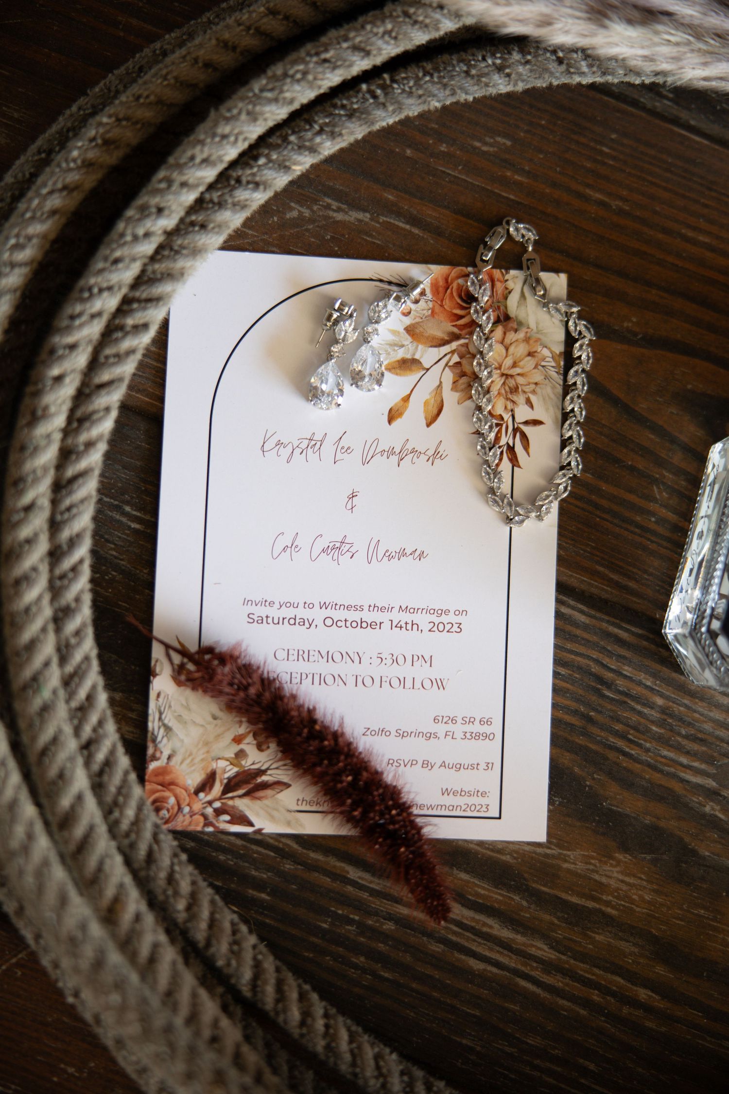 Boho wedding invitation with floral arch design, rings, and bracelet on rustic wooden surface with rope.