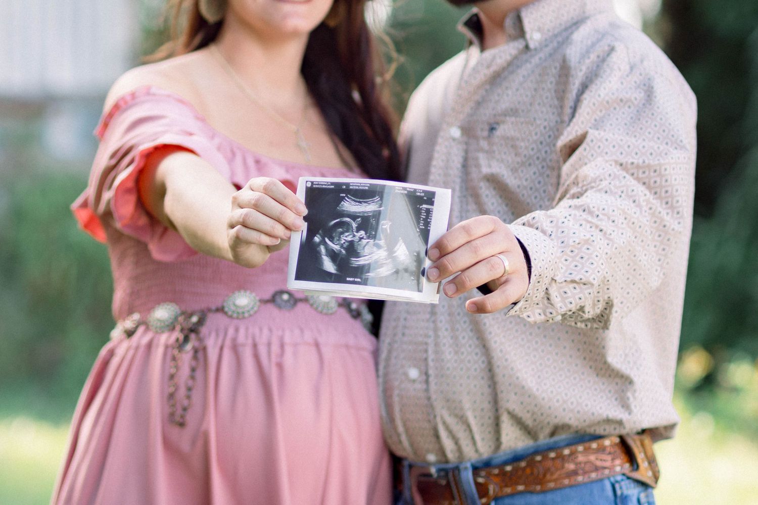 Expecting couple holding ultrasound photo outdoors, woman in pink maternity dress with cowboy partner.