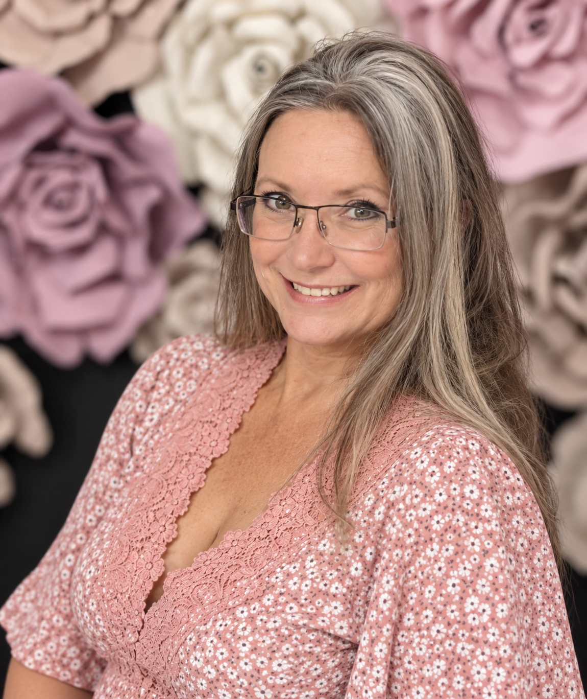 Smiling woman with gray hair and glasses wearing a pink floral top, posed in front of decorative paper roses.