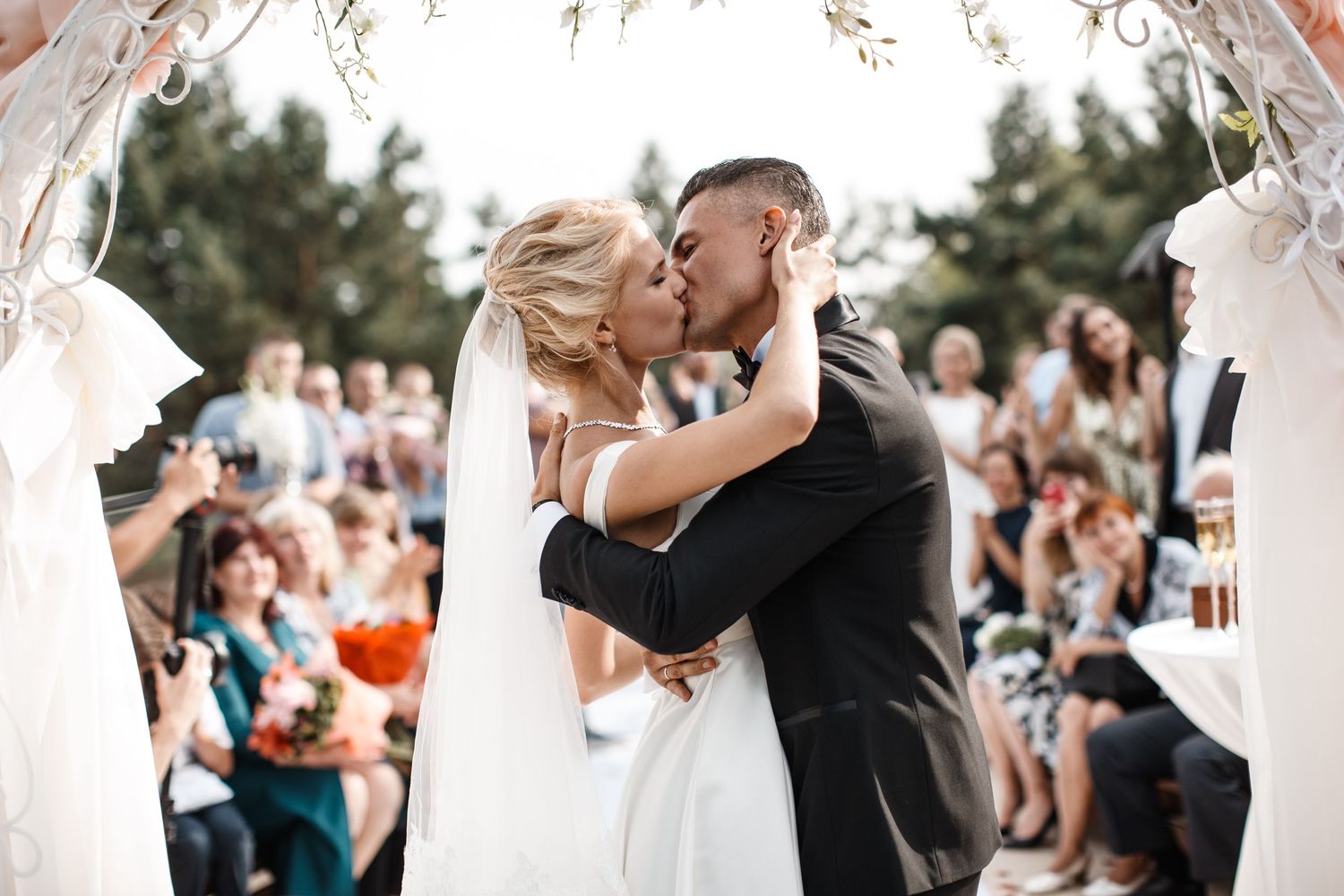 A bride and groom share their first kiss at an outdoor wedding ceremony surrounded by guests.