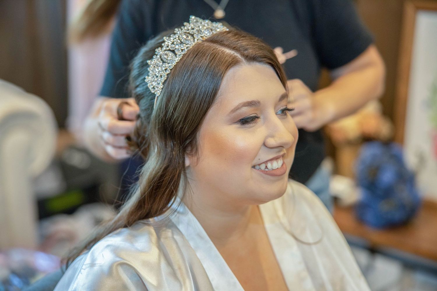 Bride smiling in satin robe wearing crystal tiara while getting hair styled for wedding day.