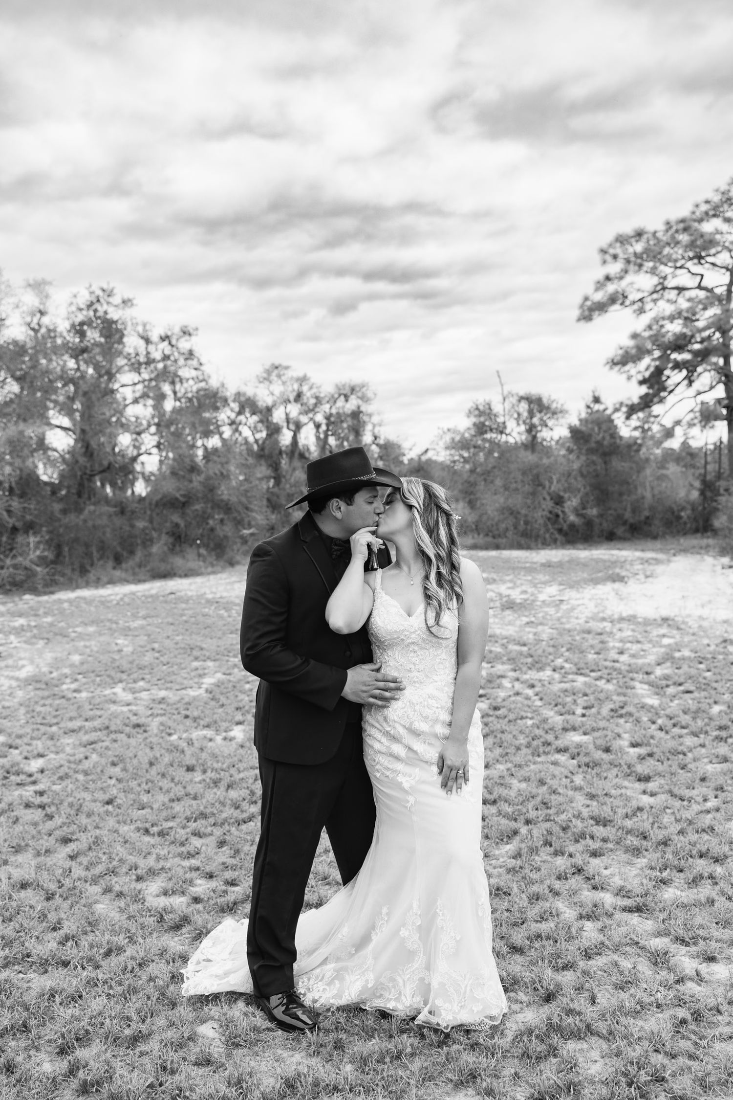 Black and white photo of bride and groom sharing a kiss outdoors, groom in black cowboy hat and suit.