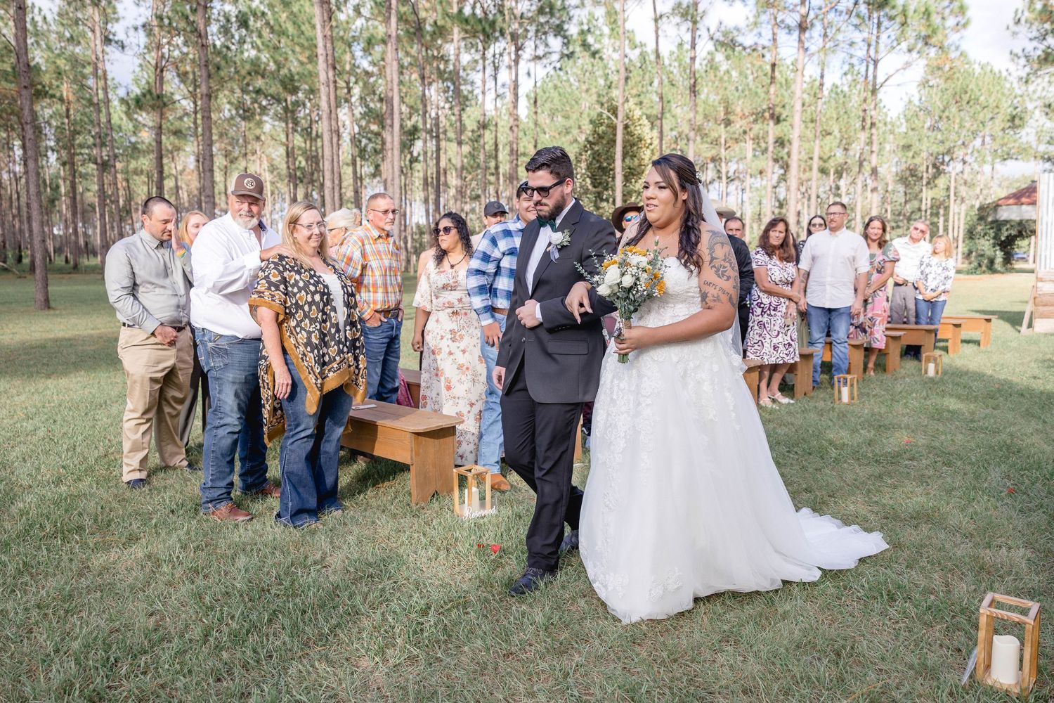 Bride in lace gown with sunflower bouquet walks down outdoor aisle with guests standing in attendance.