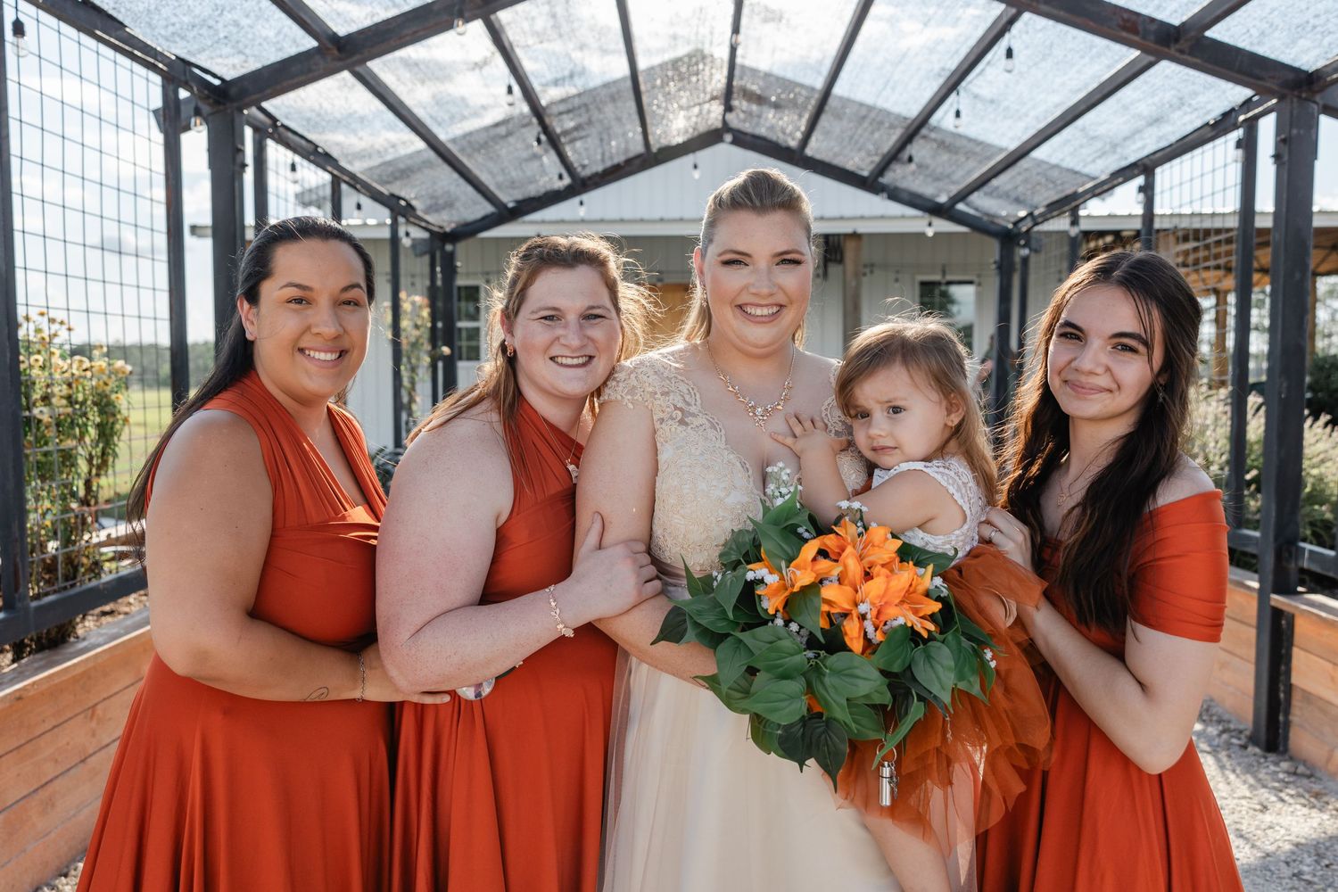 Bride with three bridesmaids in rust orange dresses and flower girl pose under greenhouse structure.