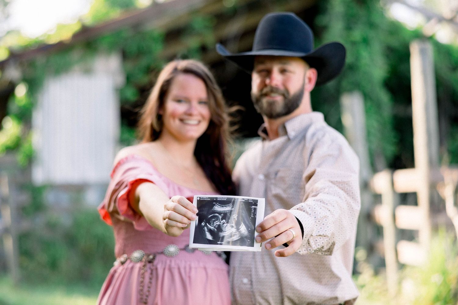 Smiling couple holding ultrasound photo for pregnancy announcement in rustic outdoor setting.