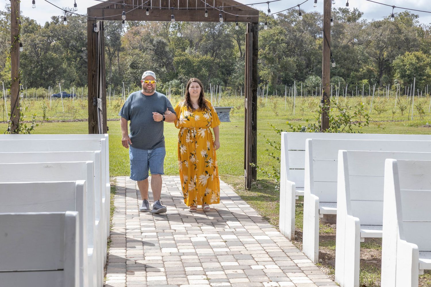 Couple walking down outdoor wedding aisle surrounded by white pews at vineyard ceremony venue.