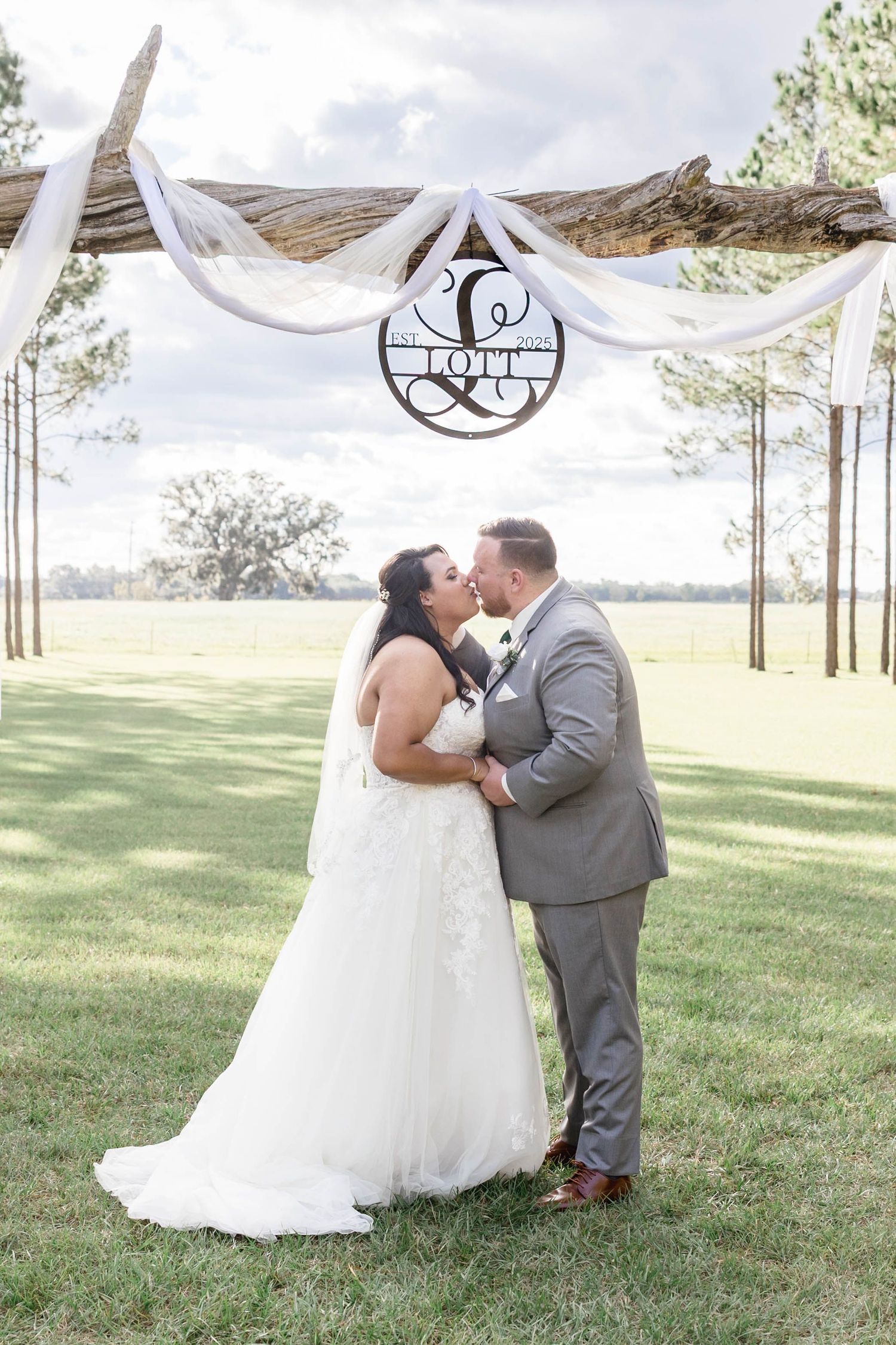 Bride and groom share first kiss under rustic wood arch with Love sign at outdoor wedding.