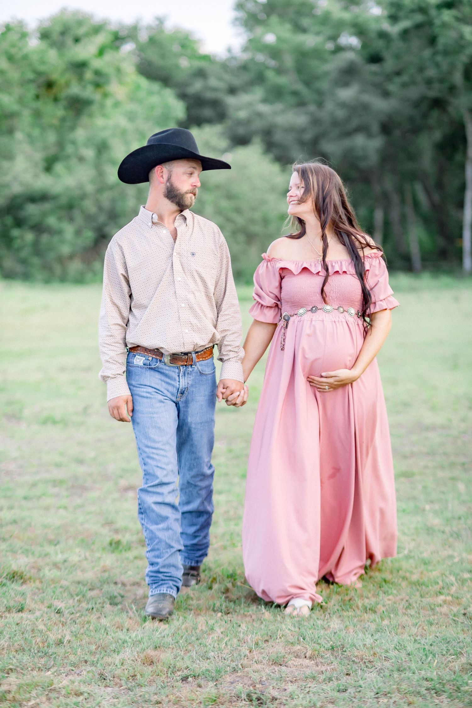 Couple walking hand in hand in green field, pregnant woman in pink dress with cowboy partner.