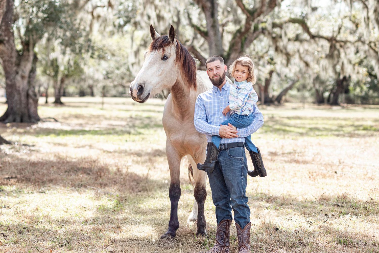 Father holding young child beside brown horse under Spanish moss-draped oak trees outdoors.