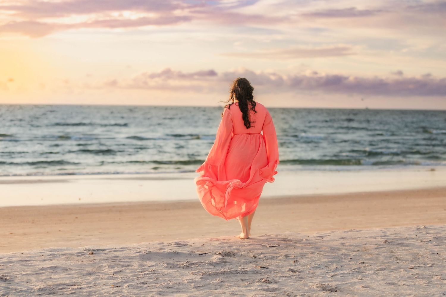 Woman in flowing coral dress walking barefoot on sandy beach at sunset.