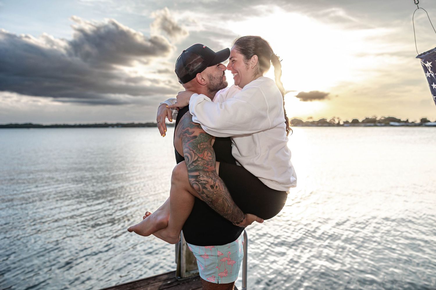 Couple sharing a romantic embrace on a dock at sunset over calm water, man with tattoos lifting woman.