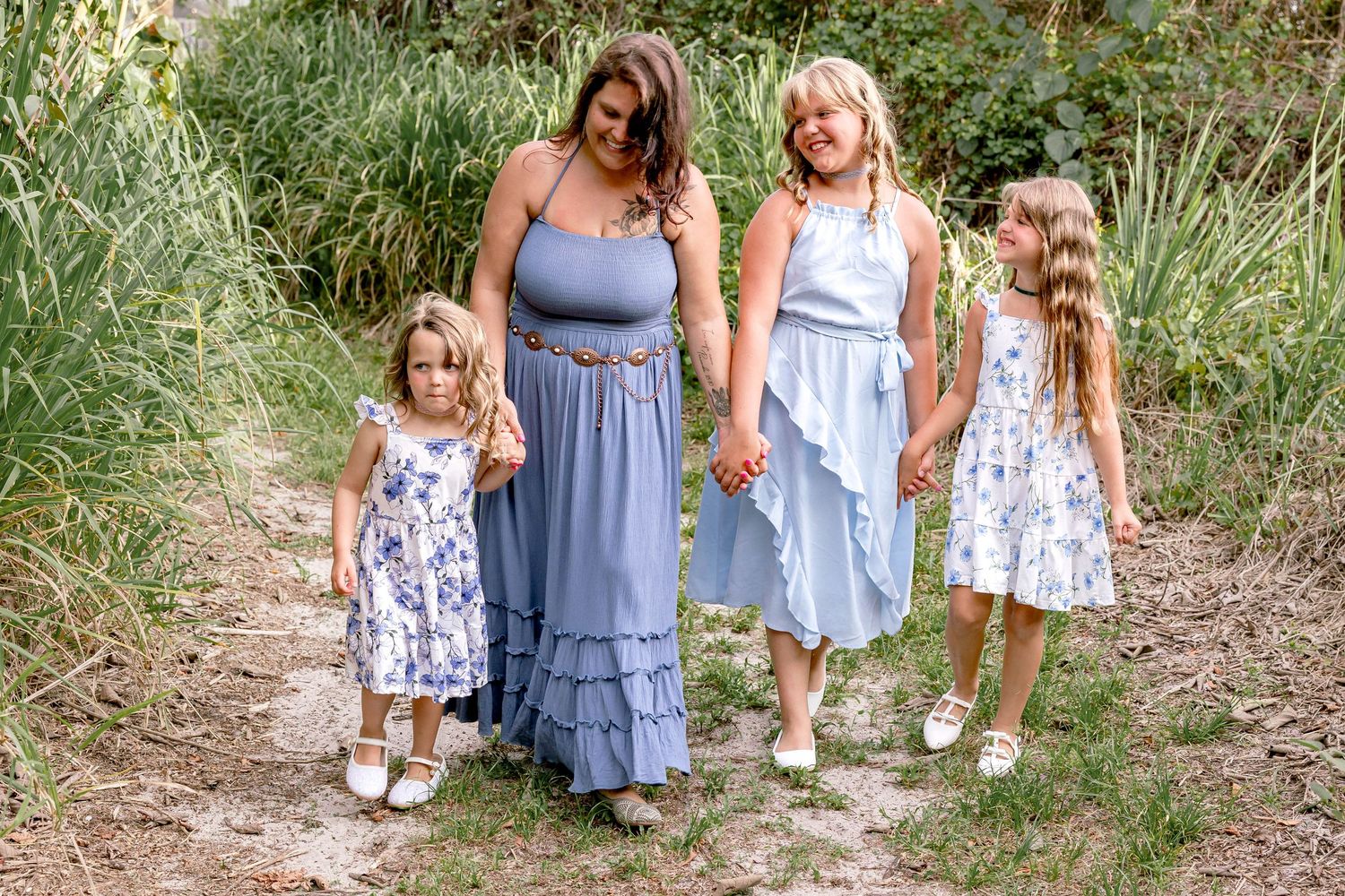 Mother and three daughters walking hand in hand on a path wearing coordinating blue and white dresses.