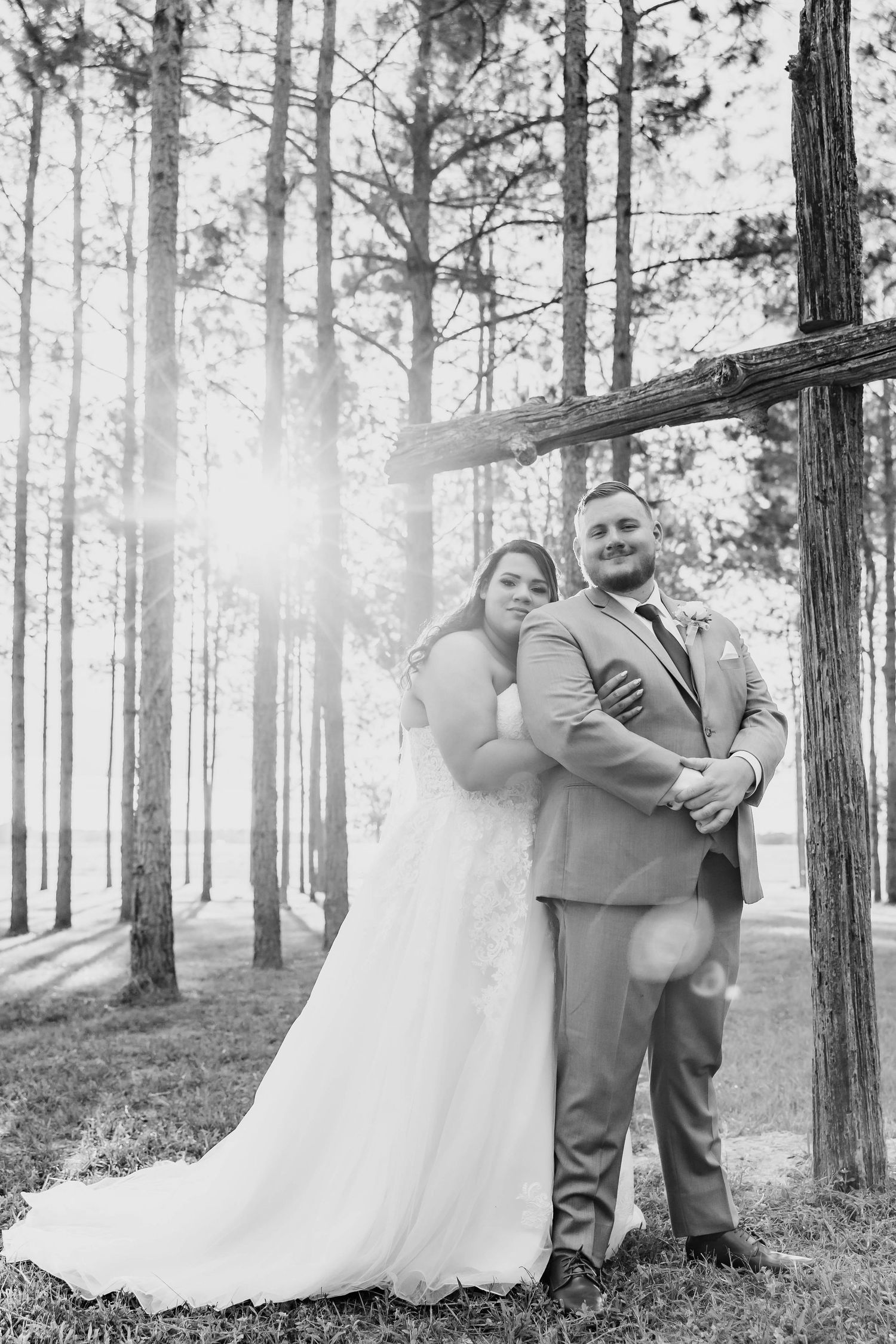 Black and white portrait of bride and groom embracing among tall pine trees with sunlight.