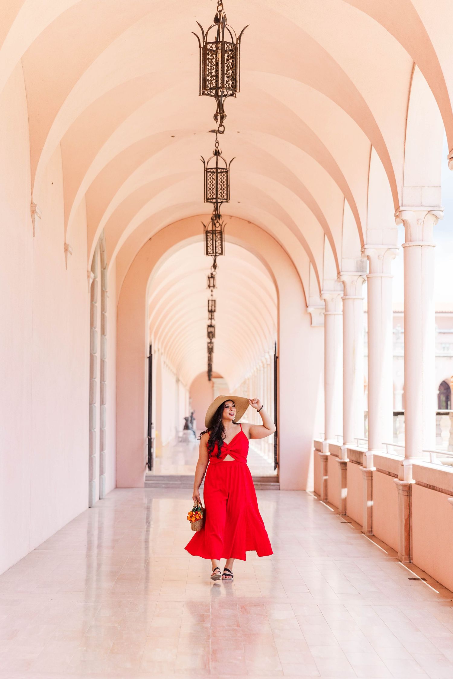 Woman in red dress and straw hat walking through pink arched colonnade corridor with hanging lanterns.