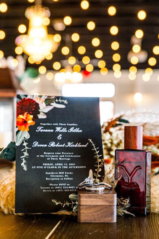 Dark floral wedding invitation with rings and red perfume bottle on wooden table.