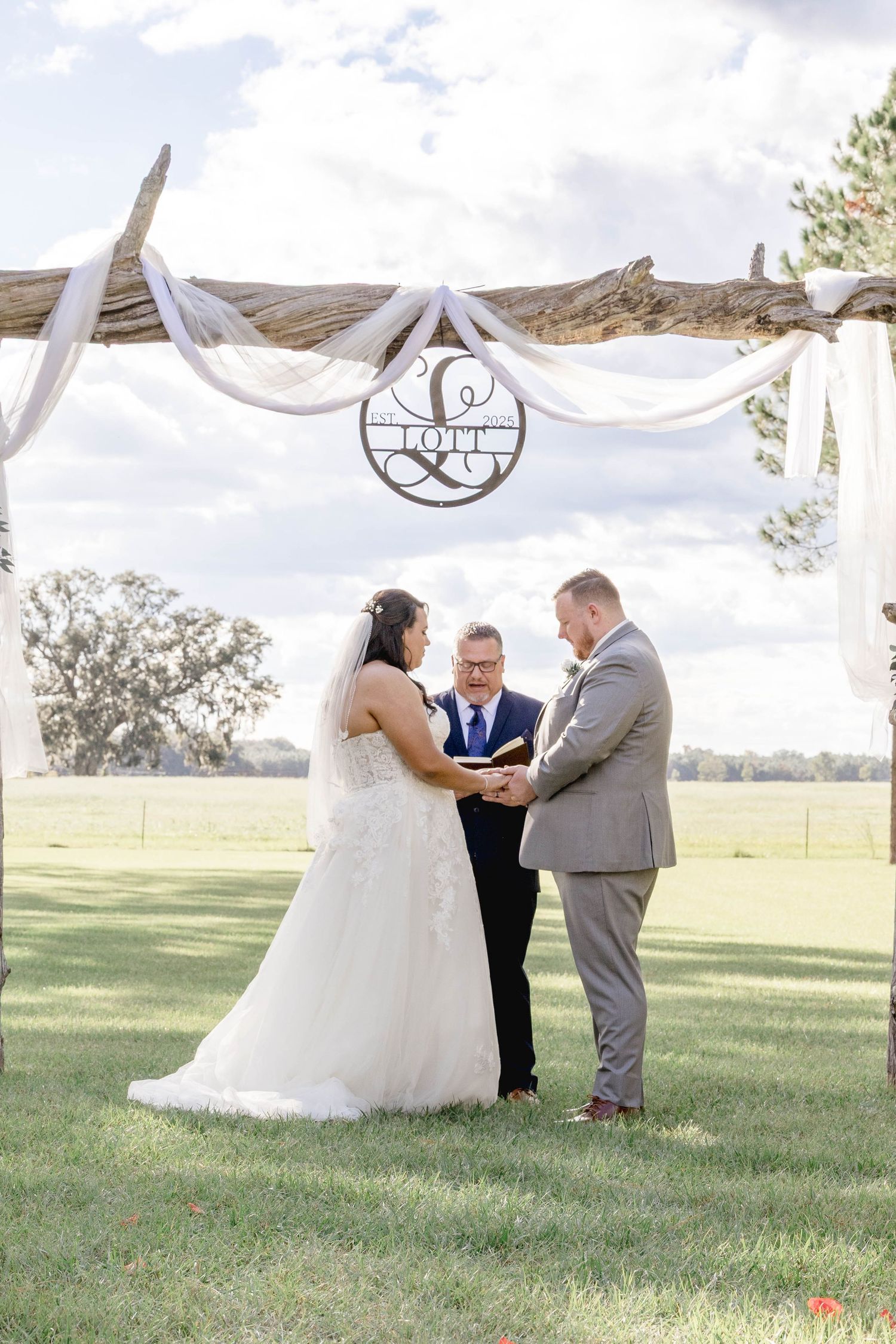 Bride and groom exchange vows under rustic driftwood arch draped with white fabric at outdoor ceremony.