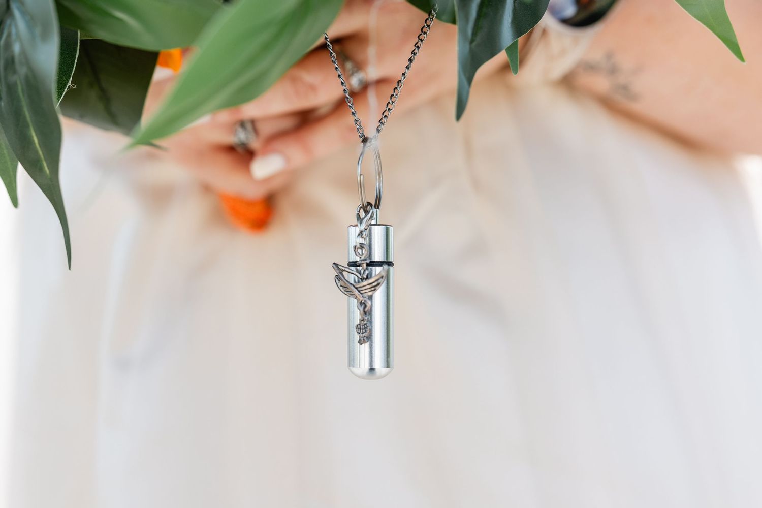 Close-up of silver cross and cylinder memorial charm necklace held against wedding bouquet greenery.