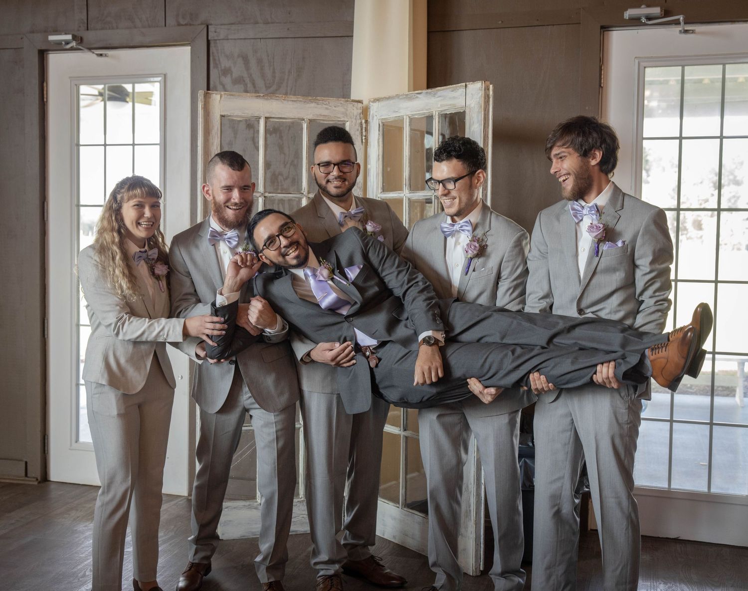 Groomsmen in gray suits playfully carrying groom while laughing together before wedding ceremony.