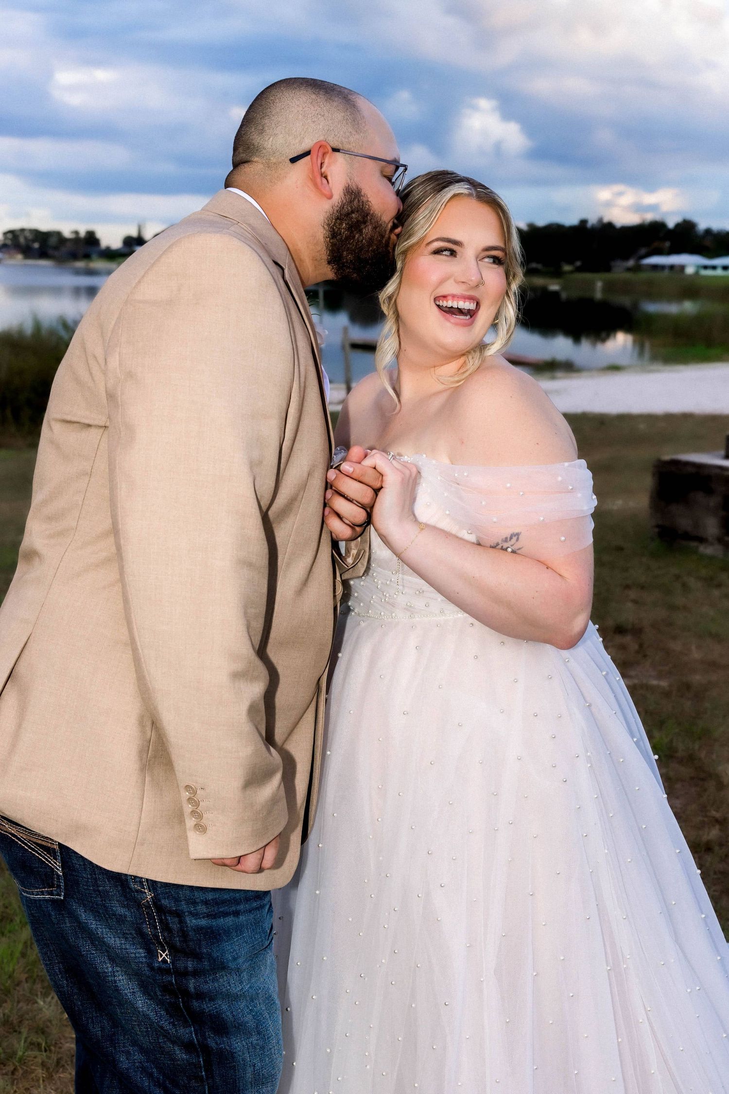 Laughing bride in off-shoulder gown holds hands with groom in tan blazer at lakeside wedding.