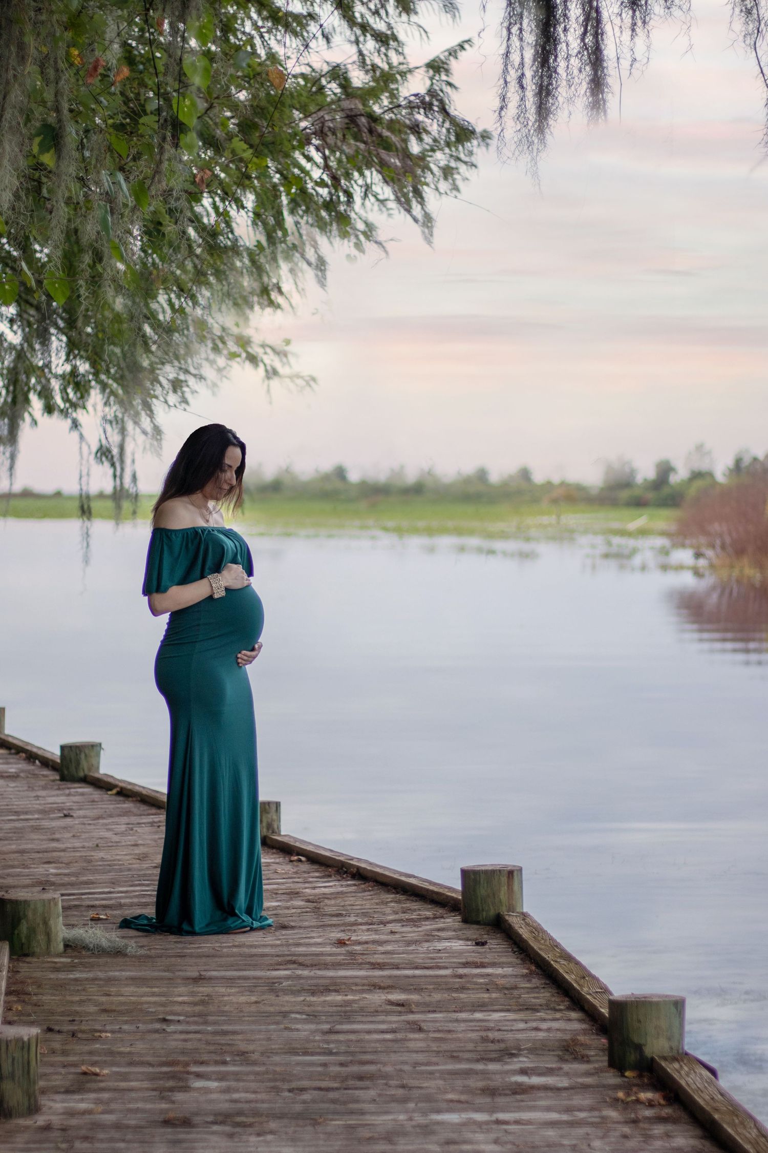 Pregnant woman in teal gown standing on wooden dock by serene lake at sunset.