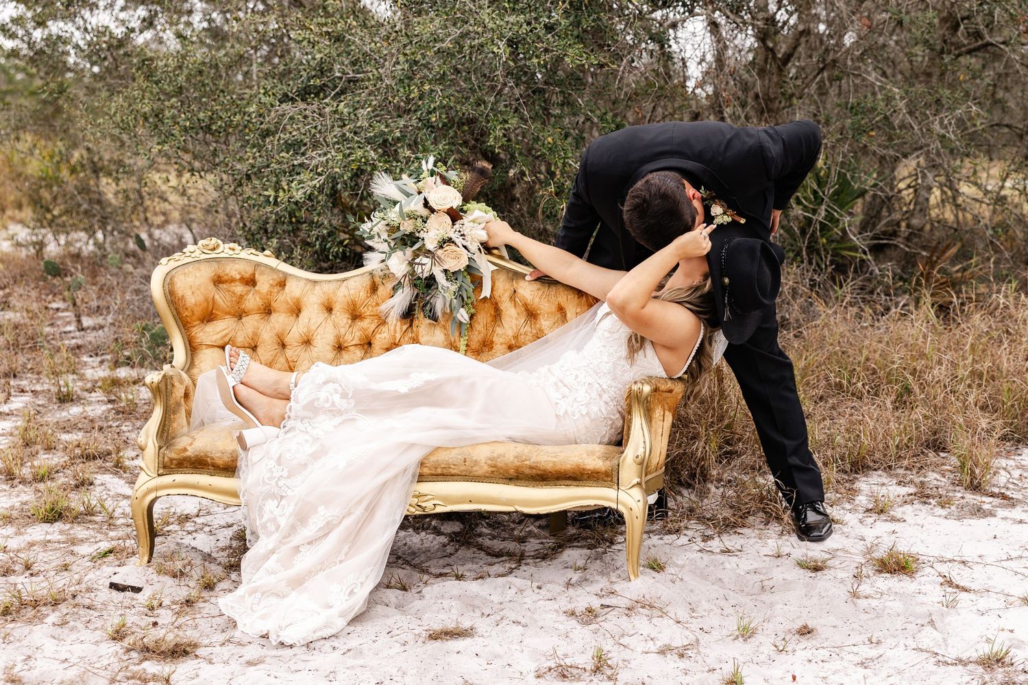 Bride in lace gown reclines on gold vintage sofa as groom leans in for a kiss outdoors.