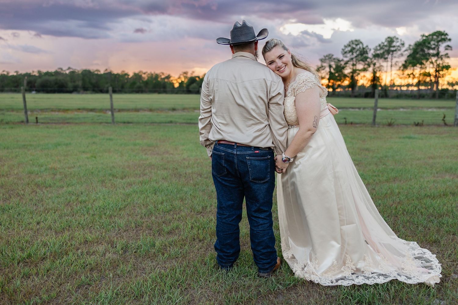 Bride in lace gown holds groom's hand in cowboy hat at sunset on green farm field.