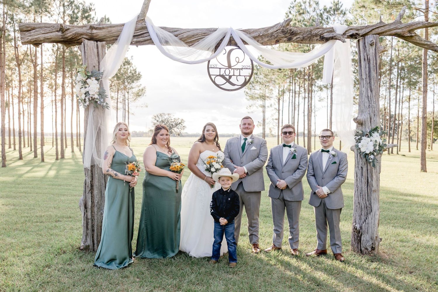 Wedding party poses under rustic wooden arch with bridesmaids in sage green gowns and groomsmen in gray suits.
