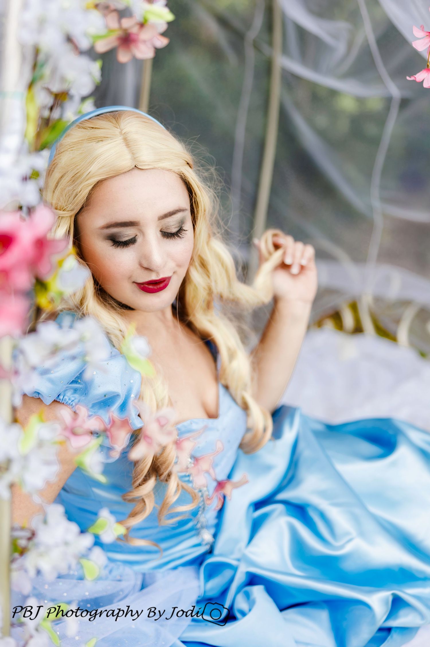 Young woman in a blue Cinderella ball gown sitting on a swing surrounded by pink flowers.