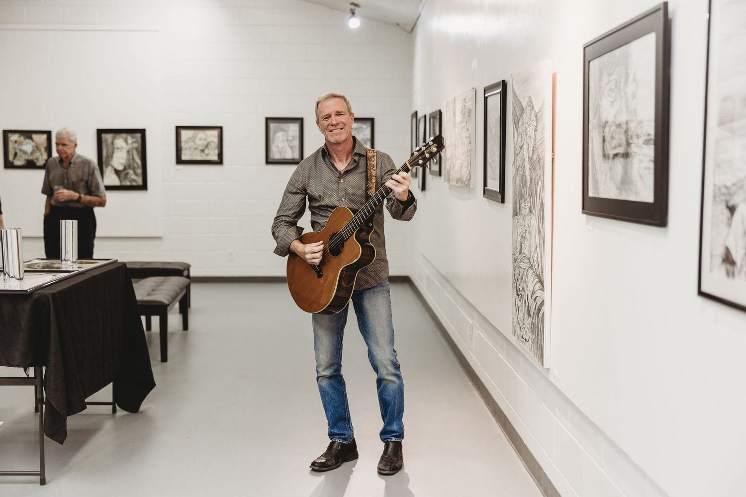 Smiling man playing acoustic guitar in white art gallery displaying framed black and white artwork.