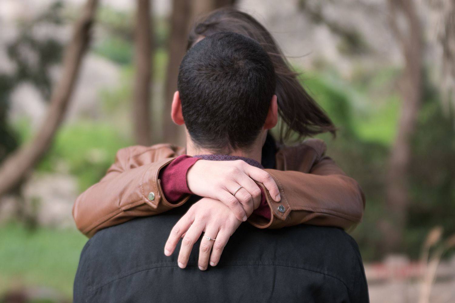 A woman with a diamond ring wraps her arms around a man from behind in a lush outdoor setting.